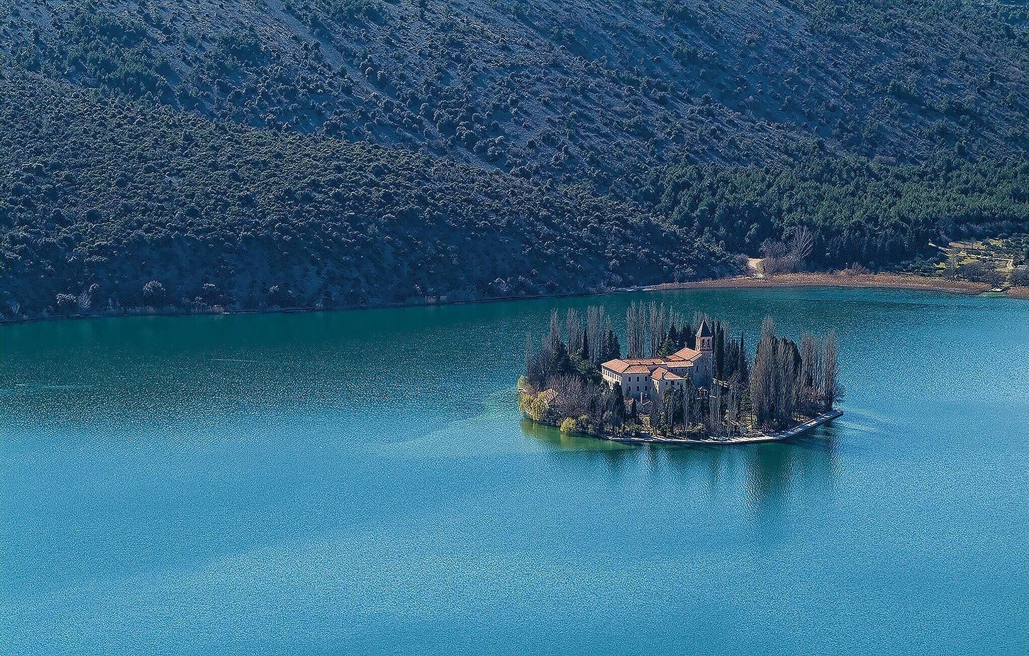 An isolated island with a building surrounded by trees, located in a large body of water with a mountainous backdrop.