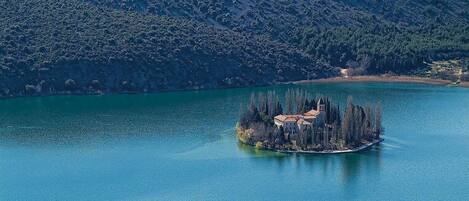 An isolated island with a building surrounded by trees, located in a large body of water with a mountainous backdrop.