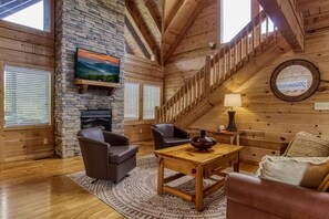 A cozy log cabin interior with a stone fireplace, wooden walls, a staircase, and a TV mounted on the wall. - Pinewood Lodge by Beyond Expectations w Hot Tub (Pigeon Forge)