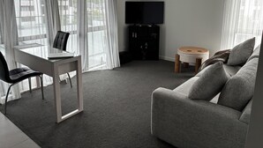 A modern living room with a grey sofa, a small round table, a glass desk with a chair, and a TV on the wall.