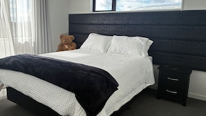 A neatly made bed with white and navy bedding, a teddy bear, and a nightstand with a power outlet.
