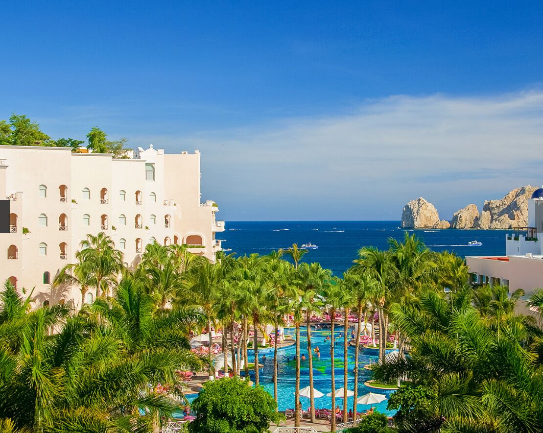 A resort with a pool surrounded by palm trees, a view of the sea, and a building with multiple balconies.