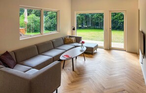 A modern living room with a grey sectional sofa, a white coffee table, and a TV on the right. Large windows provide a view of a garden. - 4 bedroom lovely home in Knebel (Knebel)