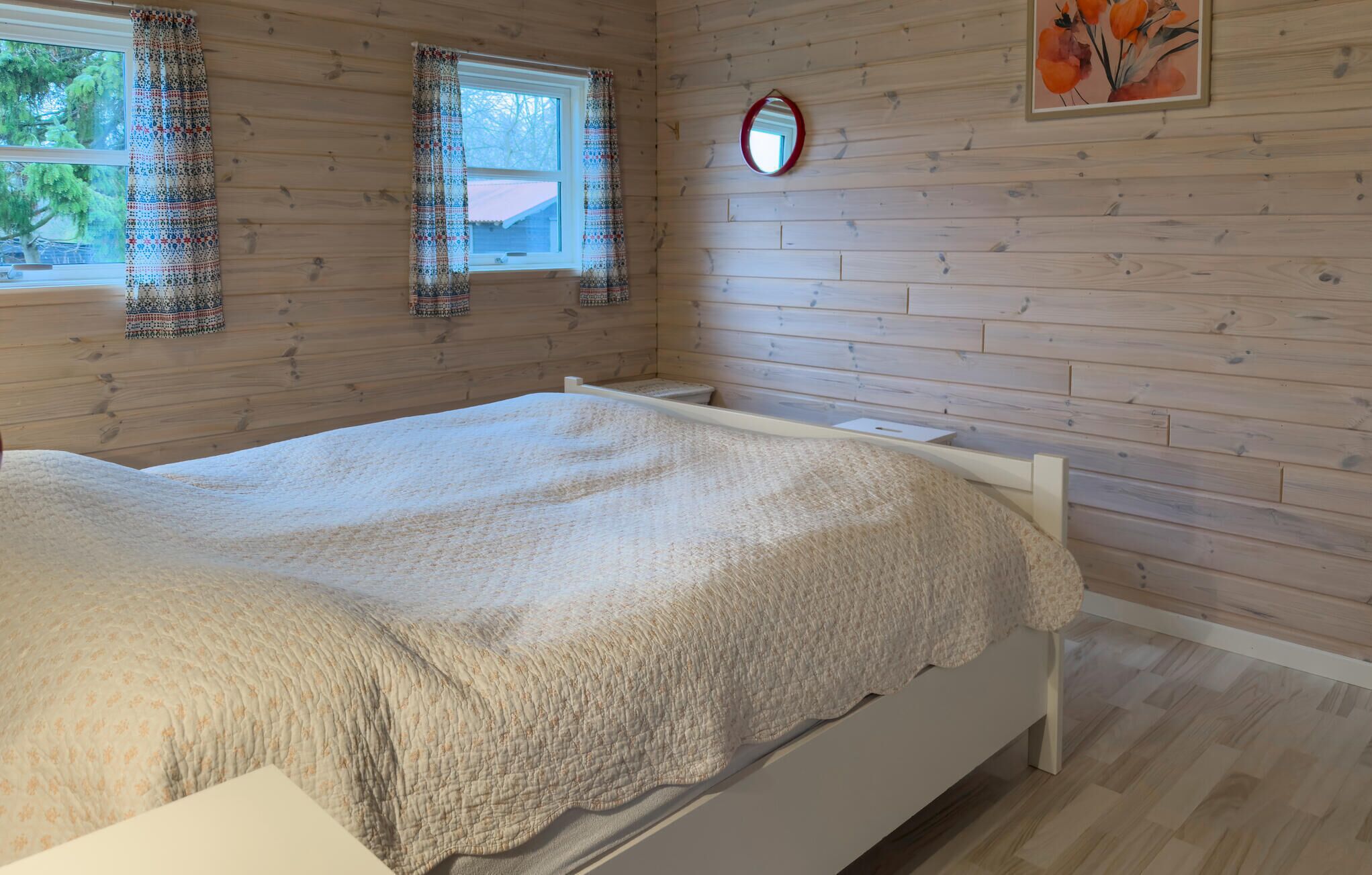A bedroom with a bed, wooden walls, a window with curtains, a mirror, and a painting.