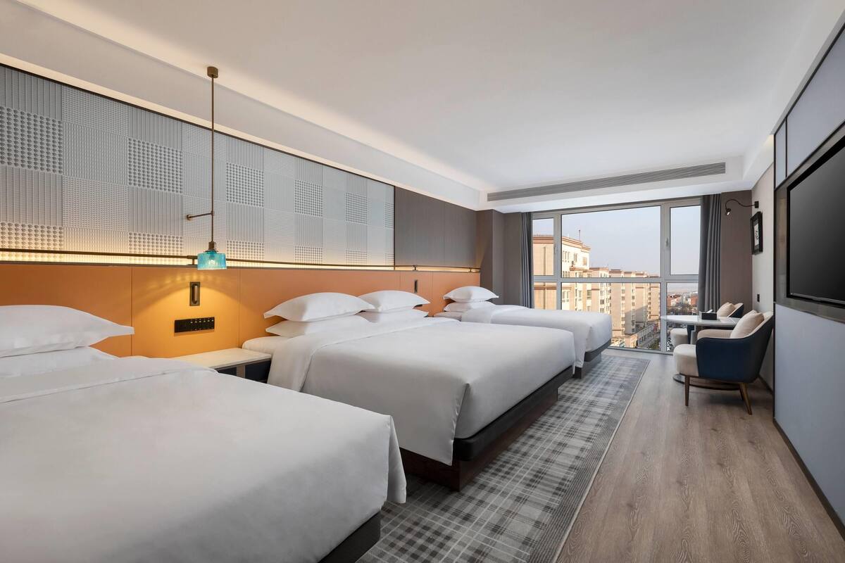 Habitación de hotel con tres camas, un escritorio, una silla y un amplio ventanal con vistas a la ciudad.