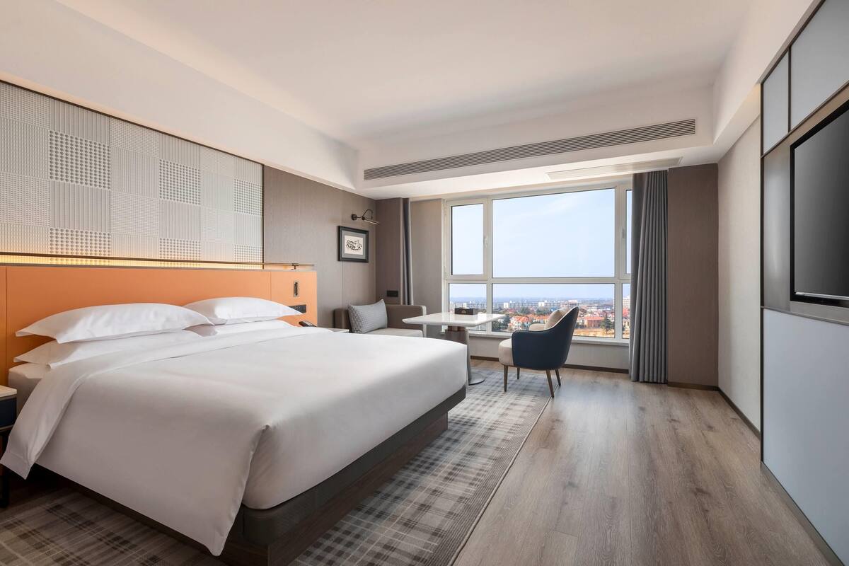 Habitación de hotel moderna con una cama grande, un escritorio, una silla y un amplio ventanal con vista a la ciudad.