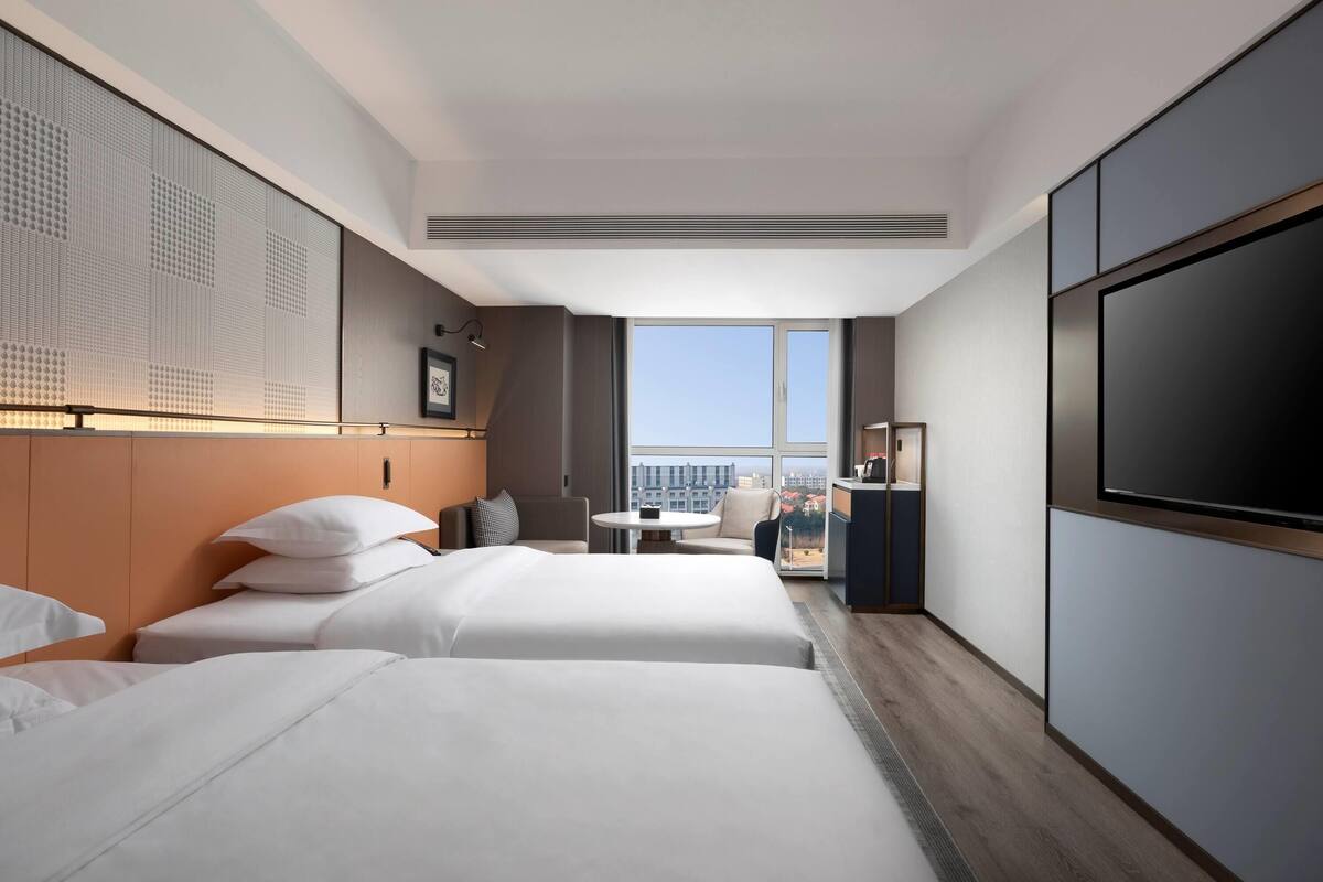 Habitación de hotel con dos camas, una mesita, una silla, televisión y un ventanal con vistas a la ciudad.