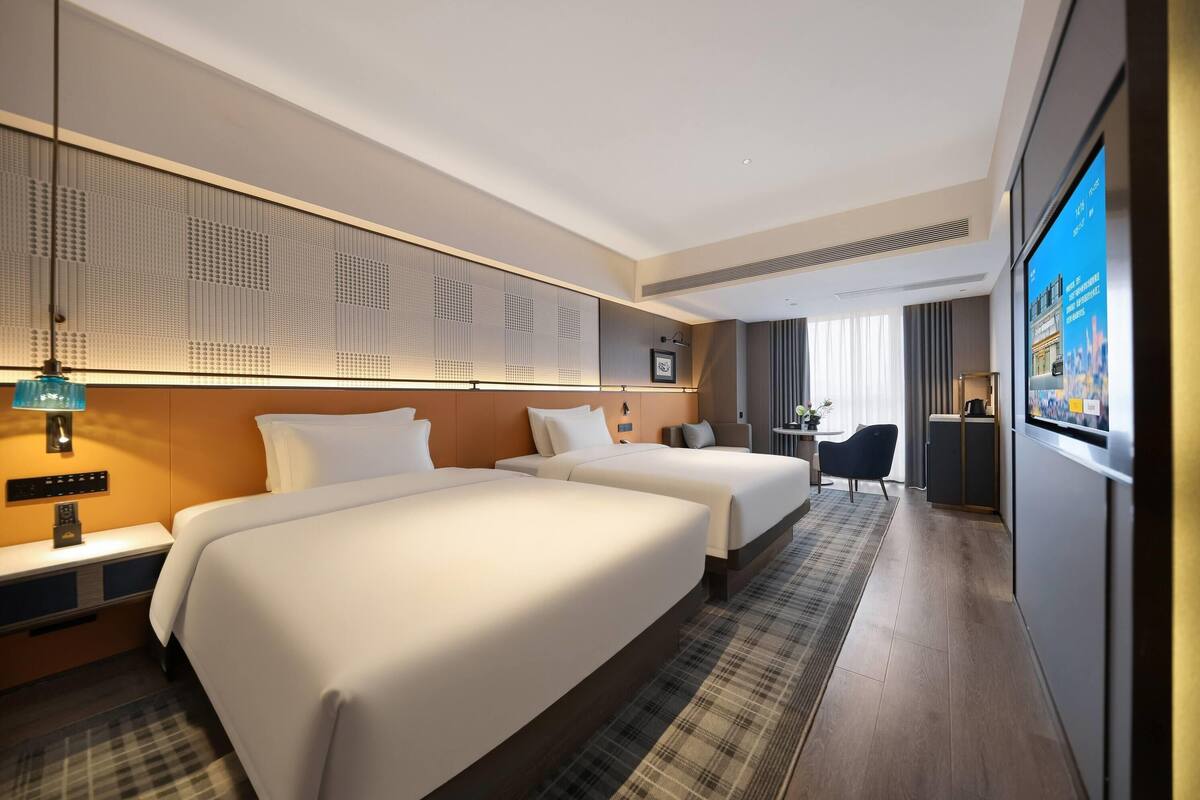 Habitación de hotel con dos camas, un escritorio, una silla, un televisor y un ventanal amplio.
