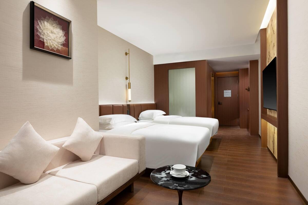 A hotel room with a sofa, two beds, a small table with a cup, a framed artwork, and a wooden floor.