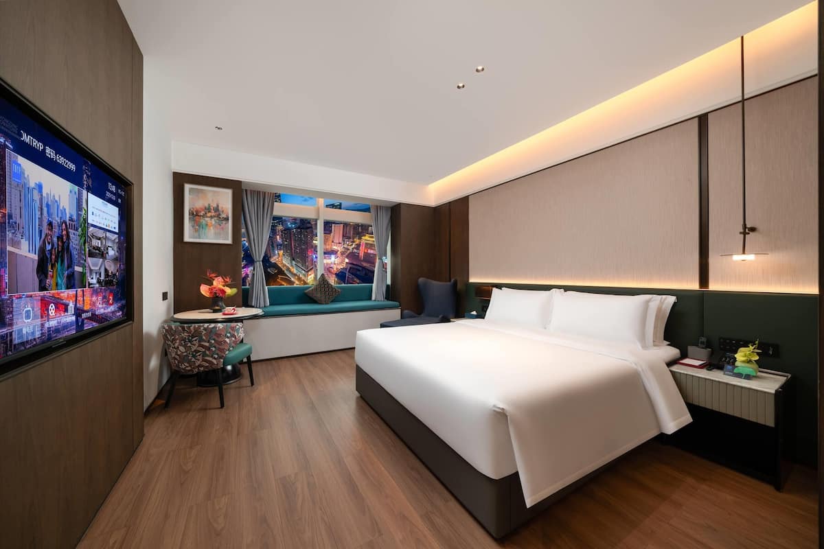 Una habitación de hotel moderna con una cama grande, una mesita con una silla, un ventanal con vistas a la ciudad y un televisor en la pared.