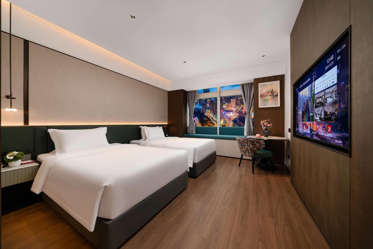 Habitación de hotel con dos camas, un escritorio, una silla, un televisor y un ventanal con vistas a la ciudad.