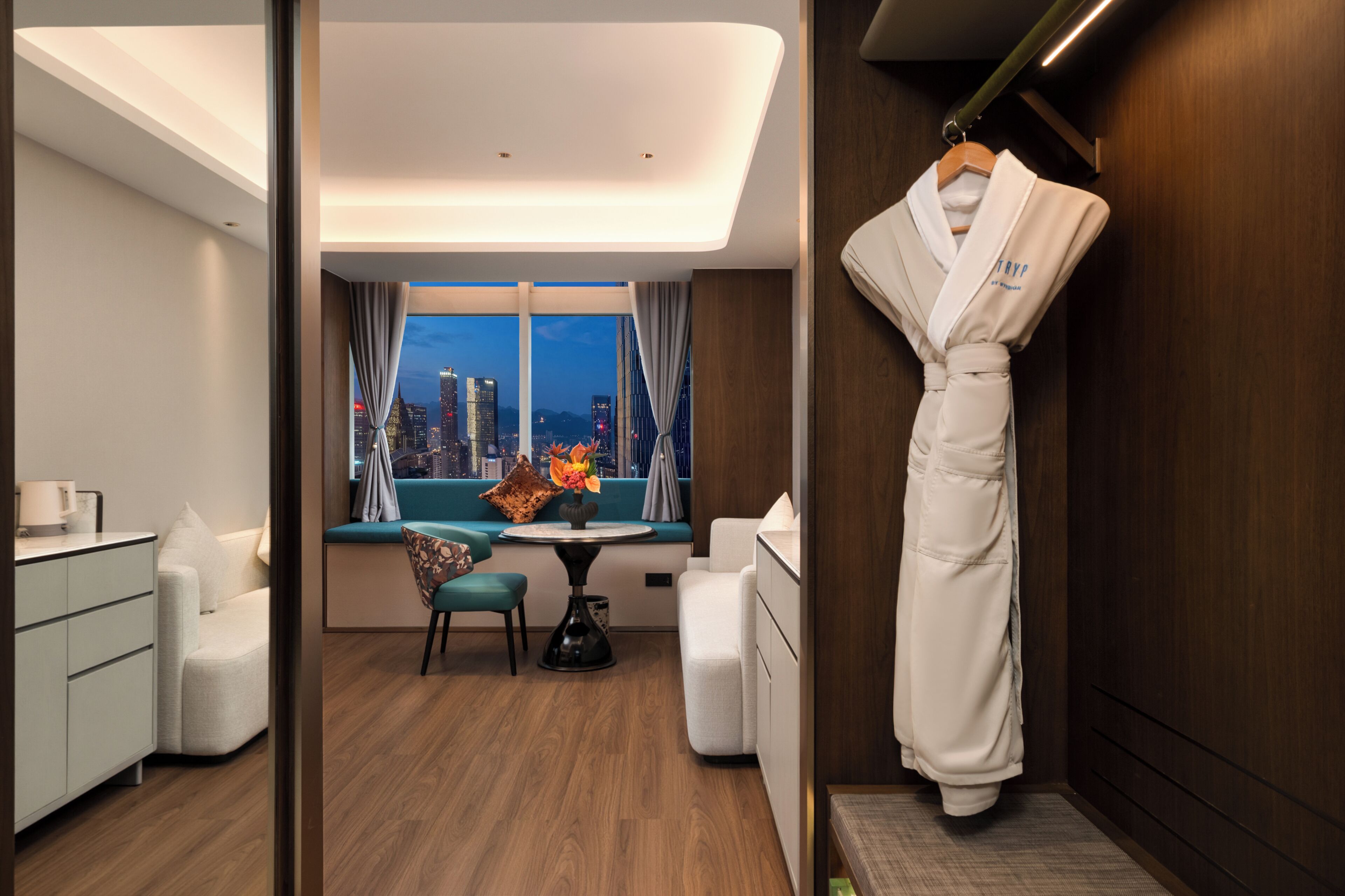 A hotel room with a city view, a dining area with a table and chairs, and a robe hanging on the wall.