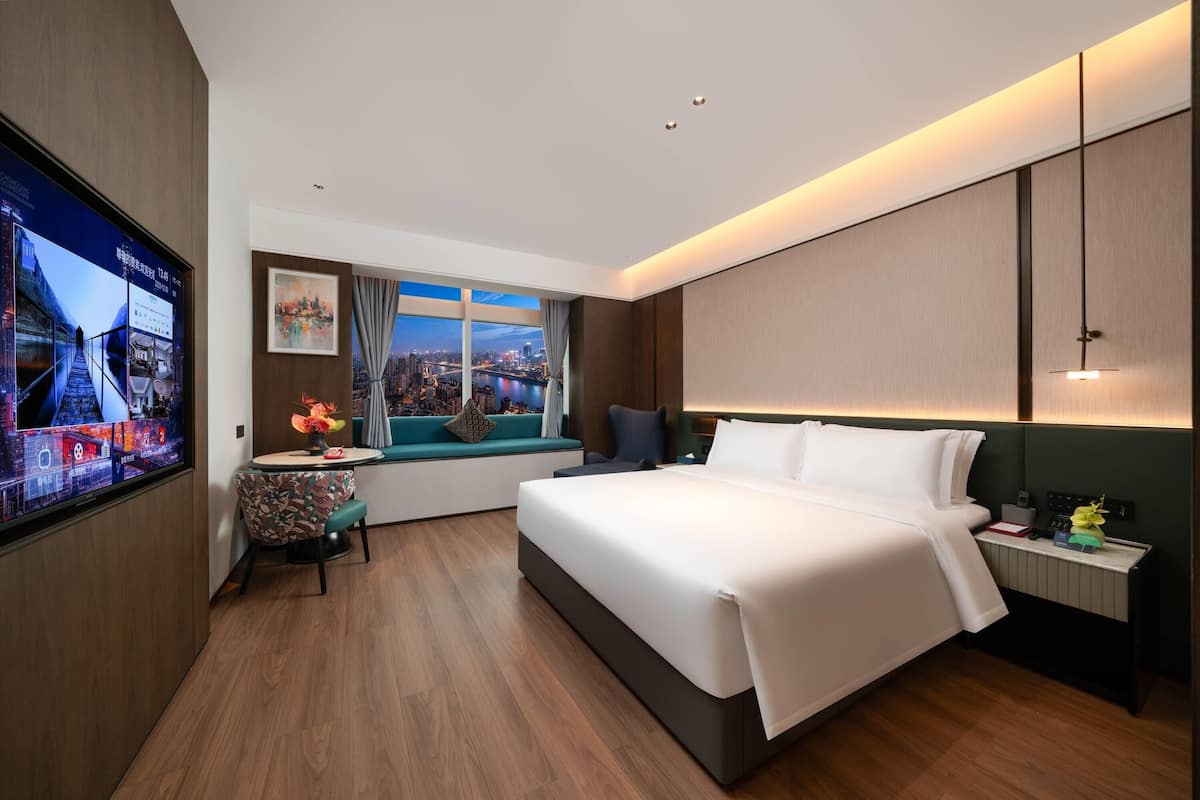 Una habitación de hotel moderna con una cama grande, un televisor montado en la pared, un ventanal con vista a la ciudad y una pequeña mesa con un jarrón.
