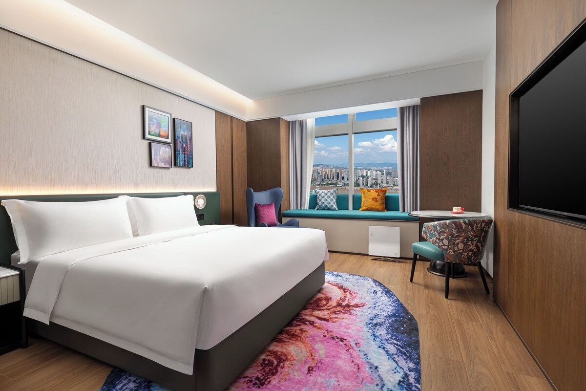Una habitación de hotel con cama, escritorio, silla, televisor y ventana con vista a la ciudad.