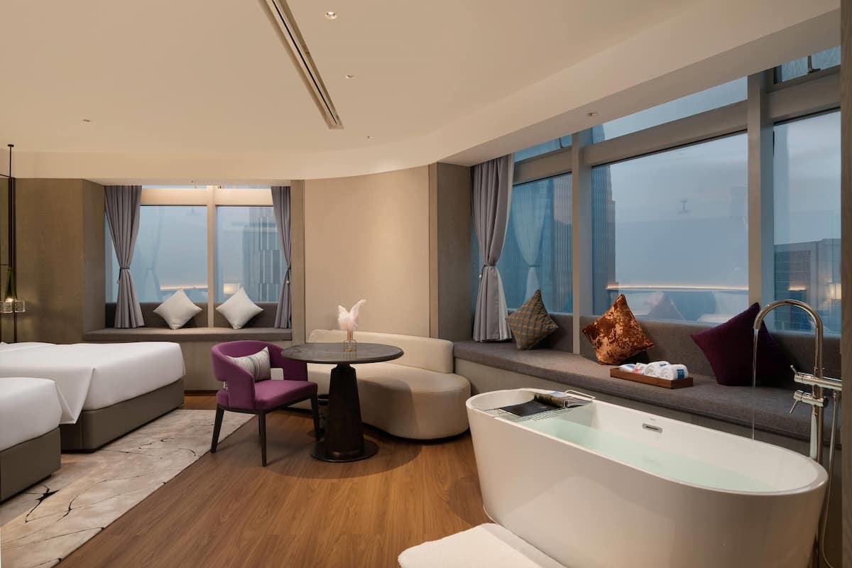 Una habitación de hotel moderna con un ventanal grande, un sofá, una mesa redonda, una bañera y una cama.