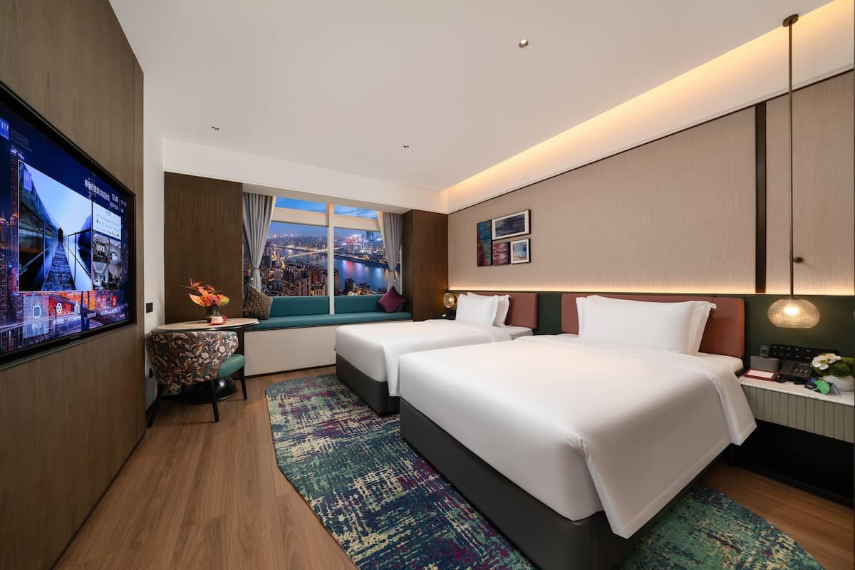 Habitación de hotel con un televisor grande, dos camas, un escritorio con lámpara y una ventana con vista a la ciudad.