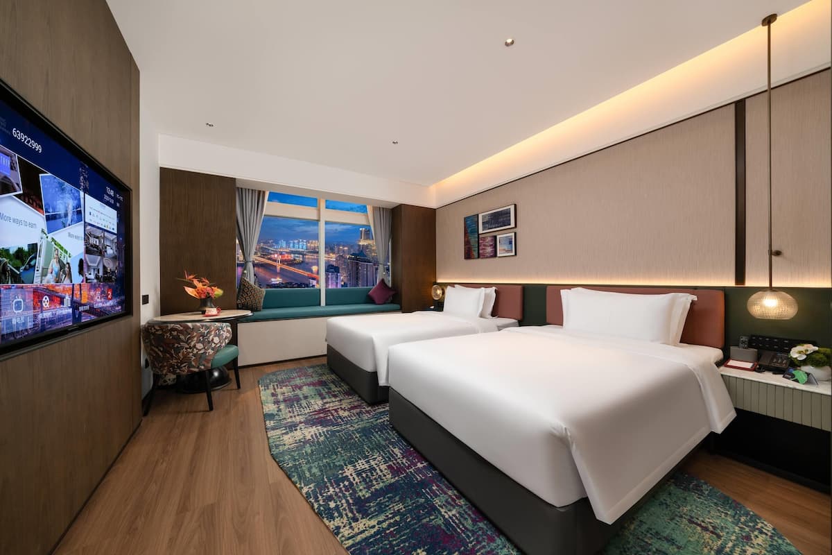 Habitación de hotel con un televisor grande en la pared, dos camas, un escritorio con lámpara y vistas a la ciudad de noche.