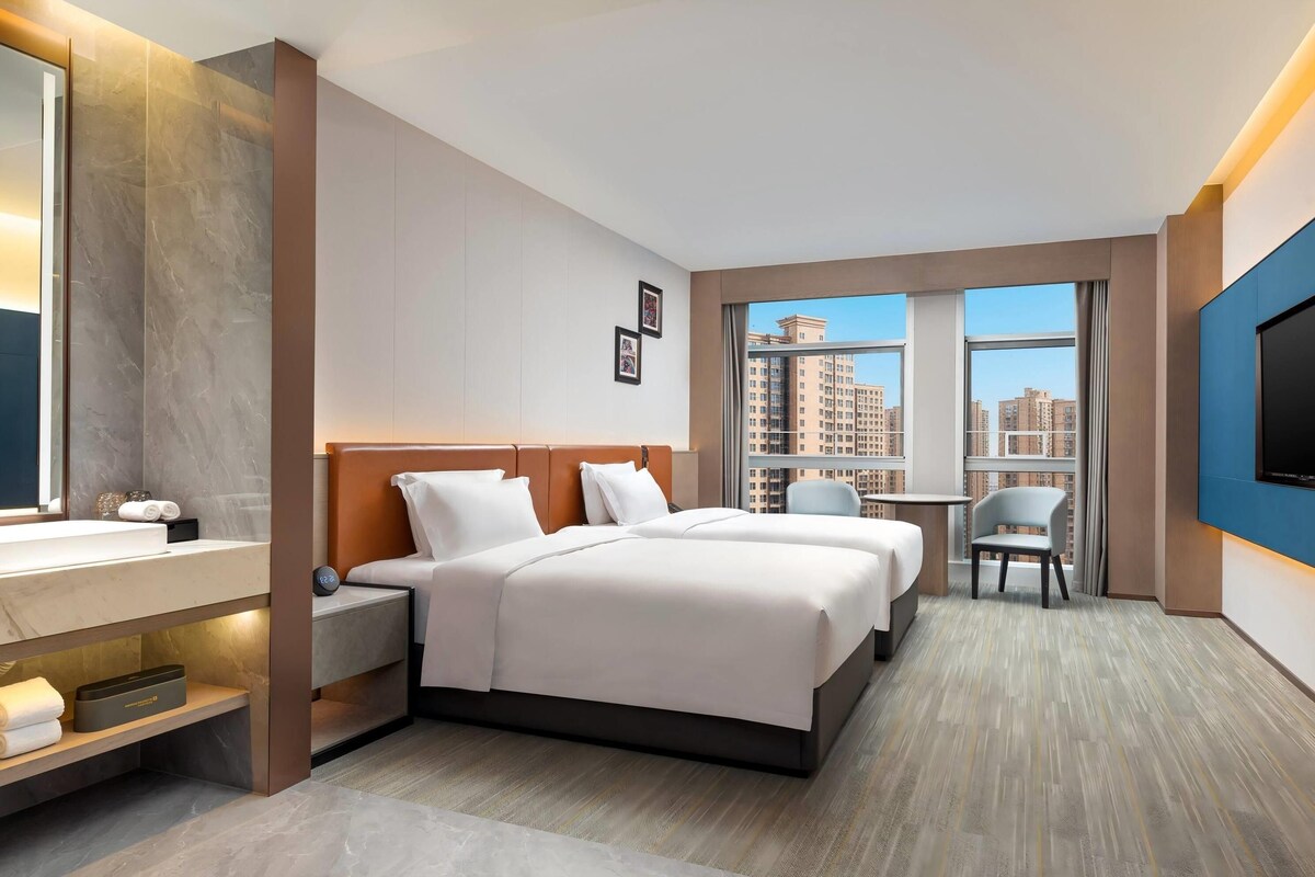 A hotel room with a bed, bedside tables, a TV, a small desk, and a window with a city view.