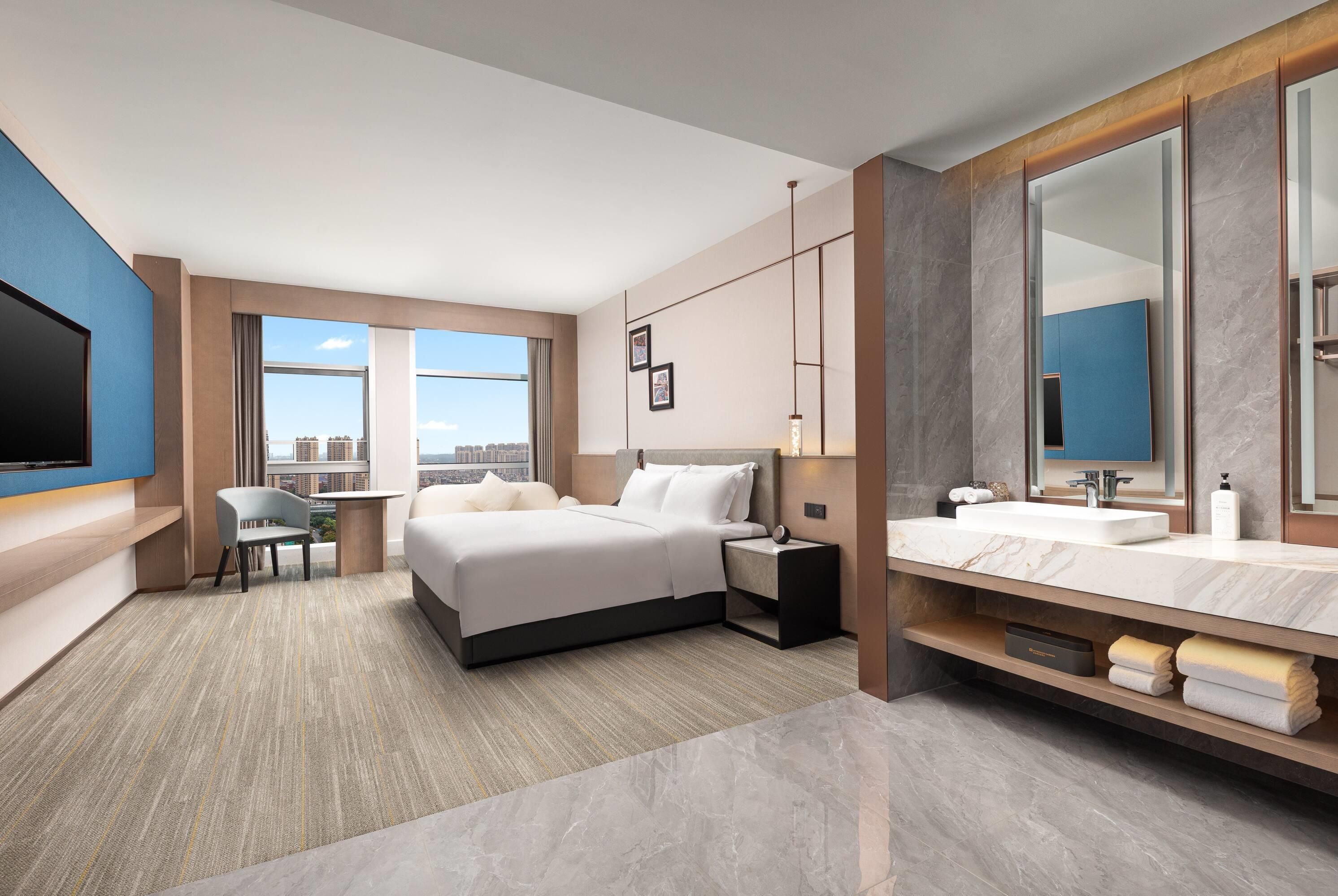 A modern hotel room with a large bed, a desk, a chair, a TV, a bathroom with a mirror and sink, and a city view.