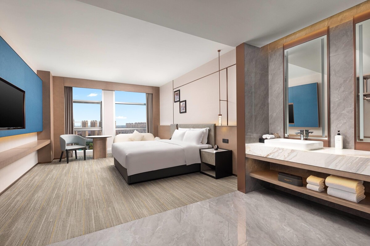A modern hotel room with a large bed, a desk, a chair, a TV, a bathroom with a mirror and sink, and a city view.