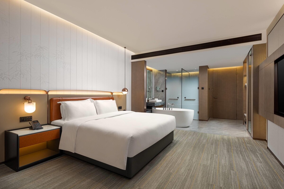 A modern hotel room with a large bed, bedside table with a phone, and a bathroom with a bathtub and shower.