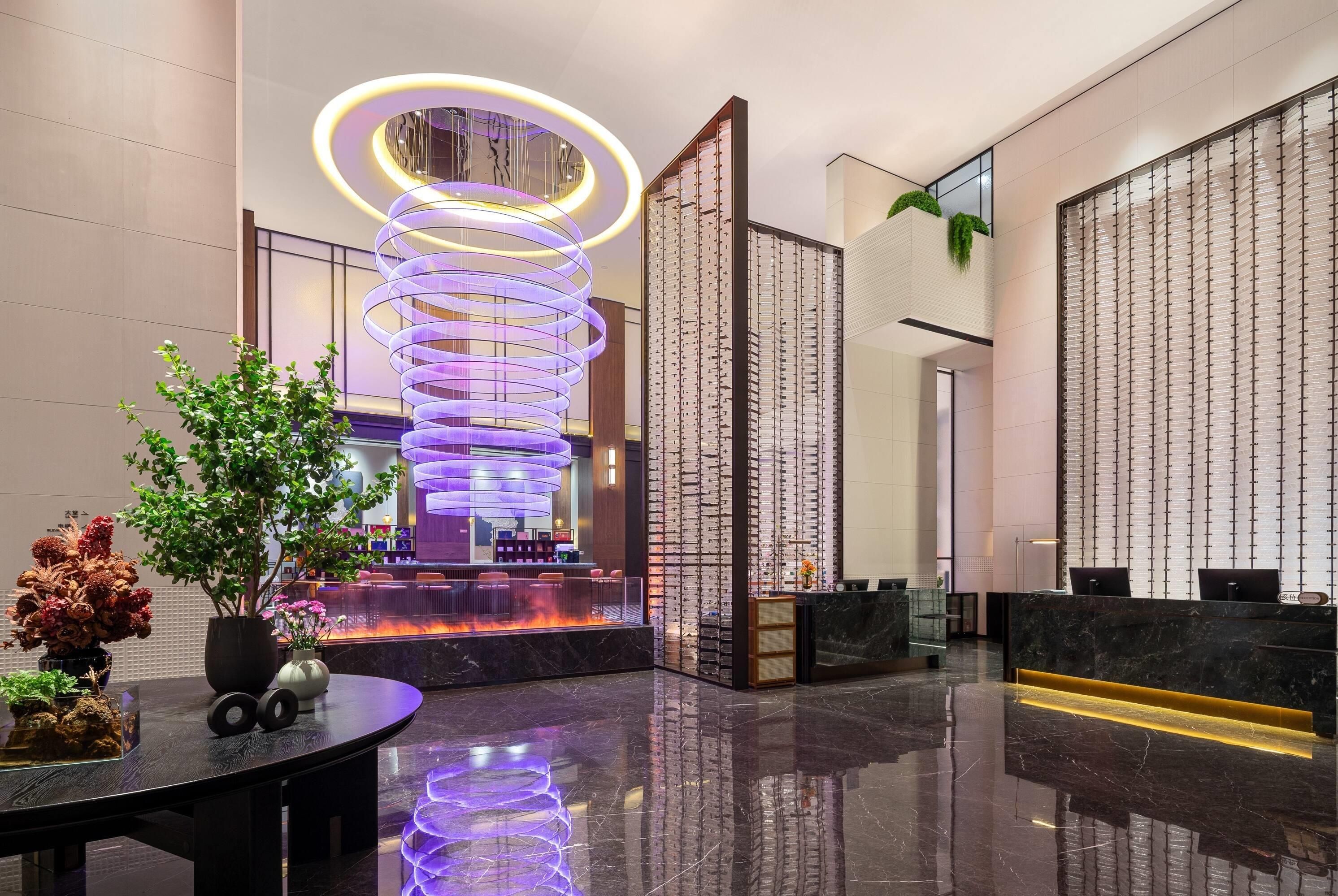 A modern lobby with a central spiral light installation, a reception desk, and a seating area with a fire feature.