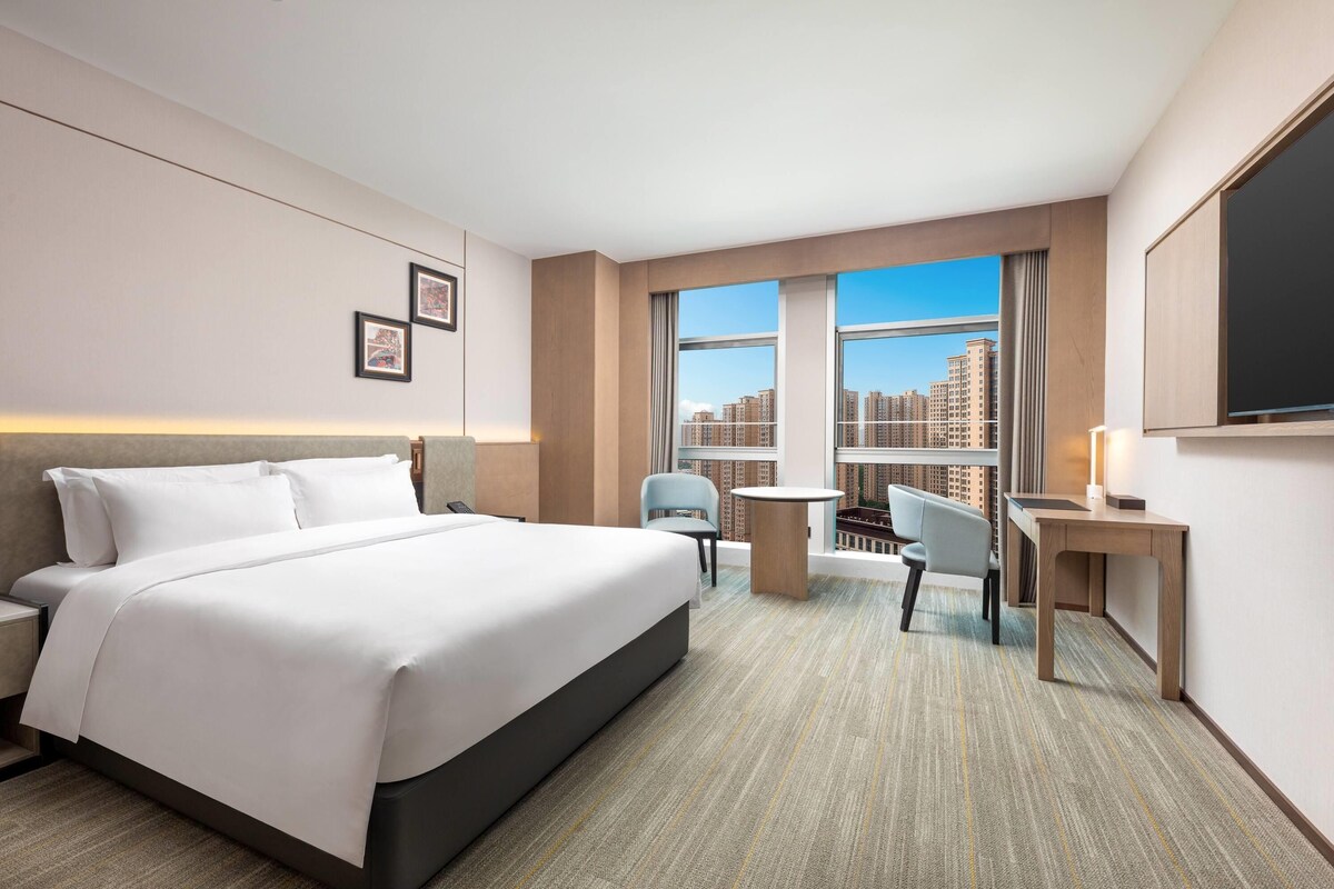 A hotel room with a large bed, a desk, a chair, and a view of a cityscape through the windows.