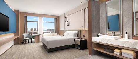 A modern hotel room with a large bed, a desk, a chair, a TV, a bathroom with a mirror and sink, and a city view.