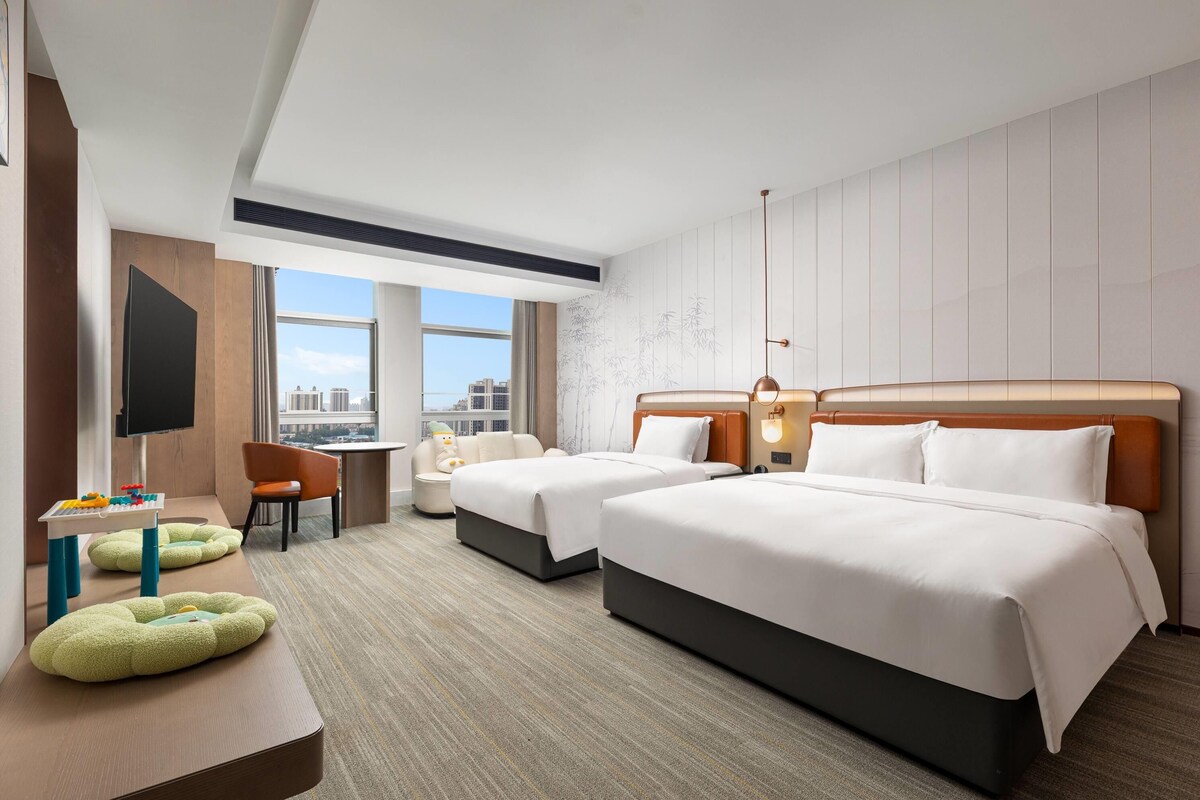 A hotel room with two beds, a desk, a chair, a television, and a view of the city.