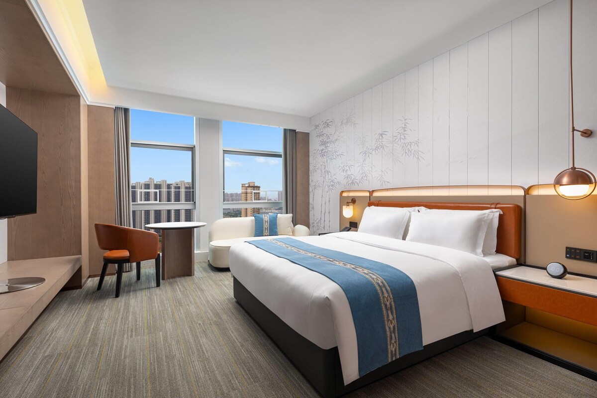 A hotel room with a bed, a desk, a chair, a TV, and a view of the city.