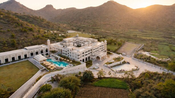 A large, white, multi-story building with a swimming pool, surrounded by a walled compound and set against a mountainous backdrop. - Wyndham Garden Udaipur Bhuwana (Udaipur)