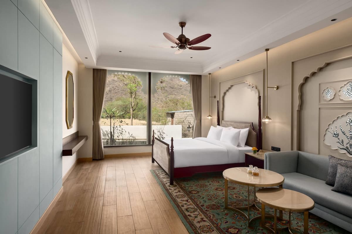 Habitación de hotel con cama, sofá, una mesita, ventilador de techo y un gran ventanal con vistas a zonas verdes.