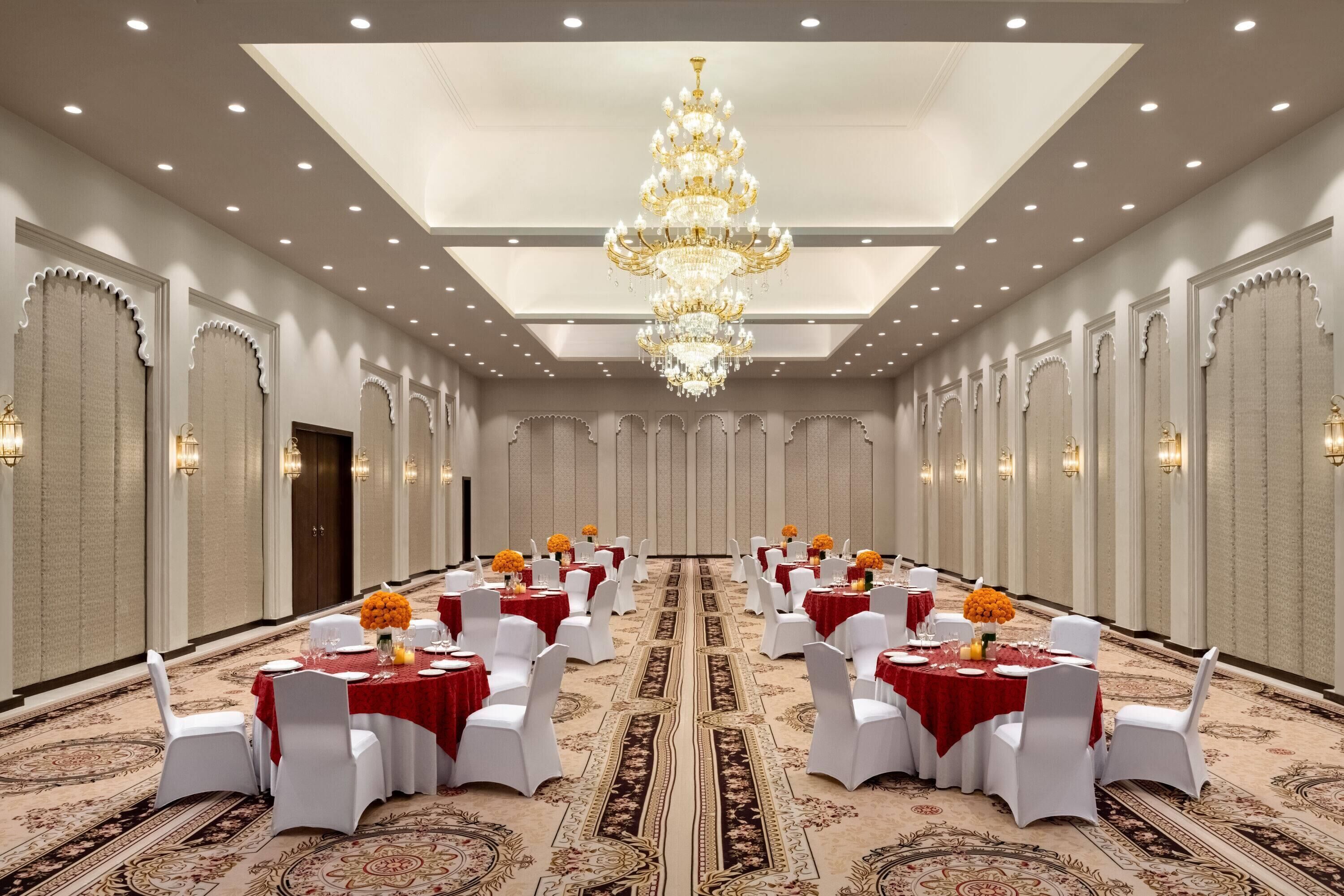 Un salón de banquetes con mesas redondas, sillas blancas, candelabros y alfombra estampada.