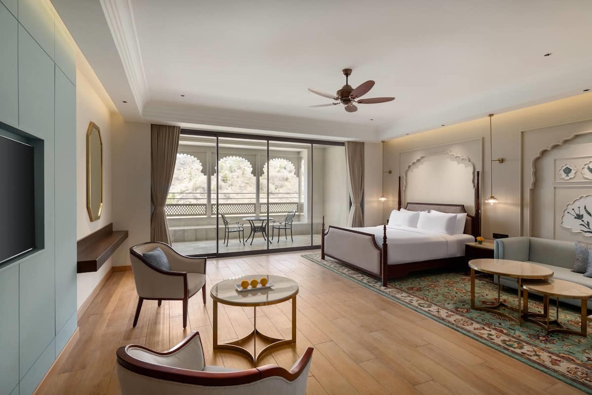 Habitación de hotel con cama, dos sofás, televisión, ventilador de techo y un gran ventanal con vistas.