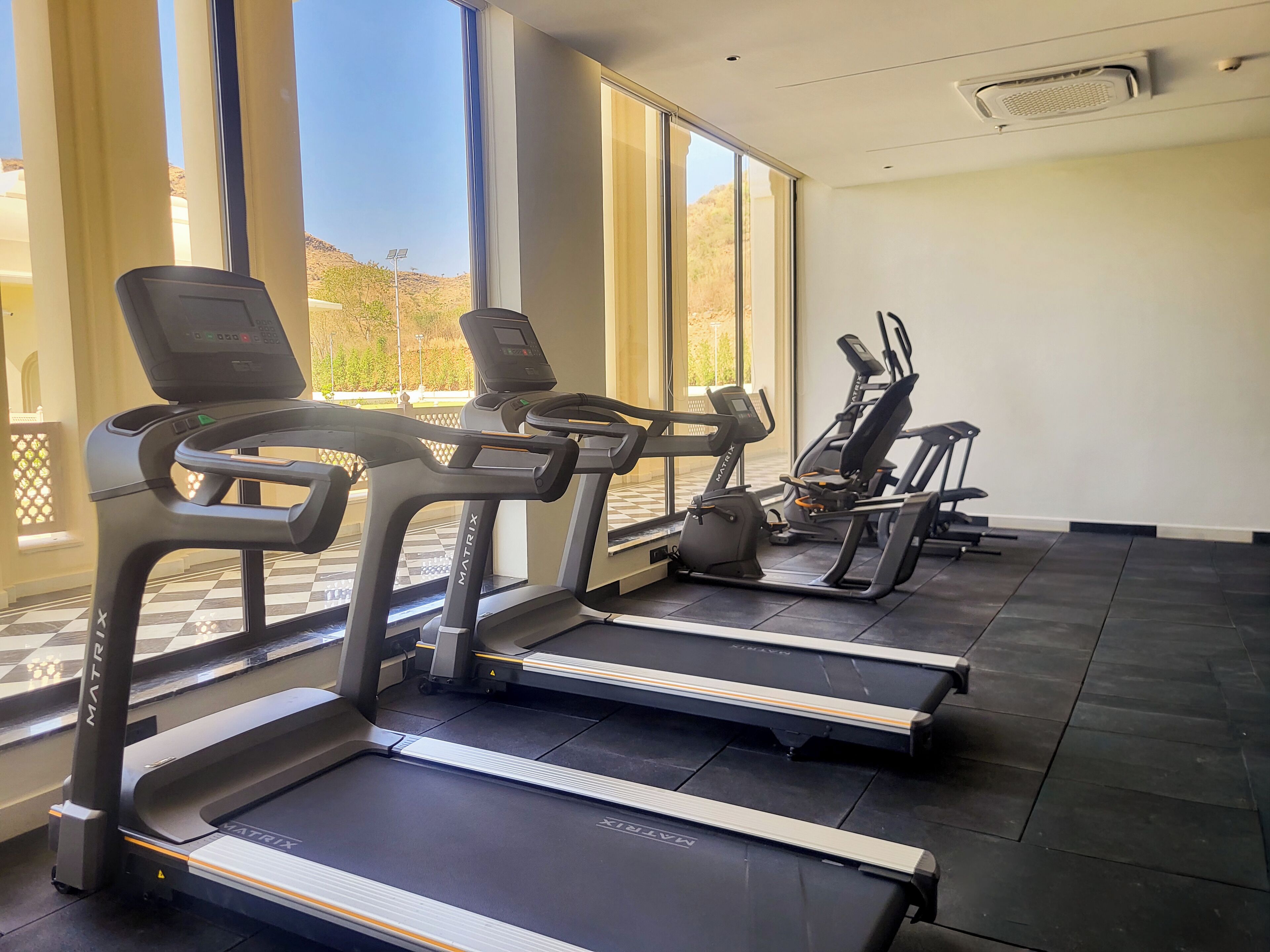 Une salle de sport équipée de tapis de course et d’appareils d’exercice, dotée de grandes fenêtres et d’un sol à carreaux.