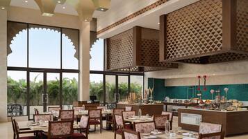 A restaurant with multiple dining tables and chairs, a buffet counter, and large windows with a view of greenery.
