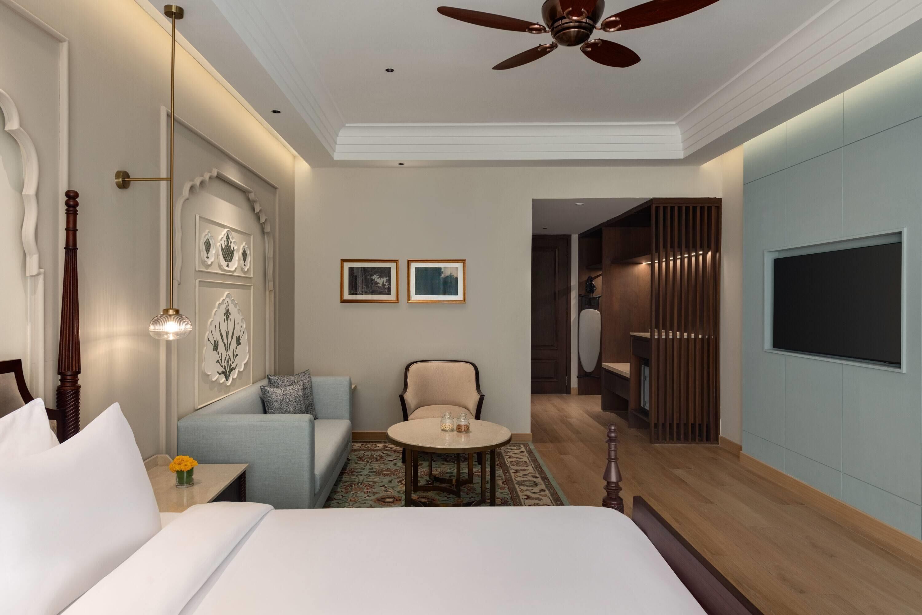 A hotel room with a bed, a sofa, a round table, a ceiling fan, a TV, and a door leading to a bathroom.