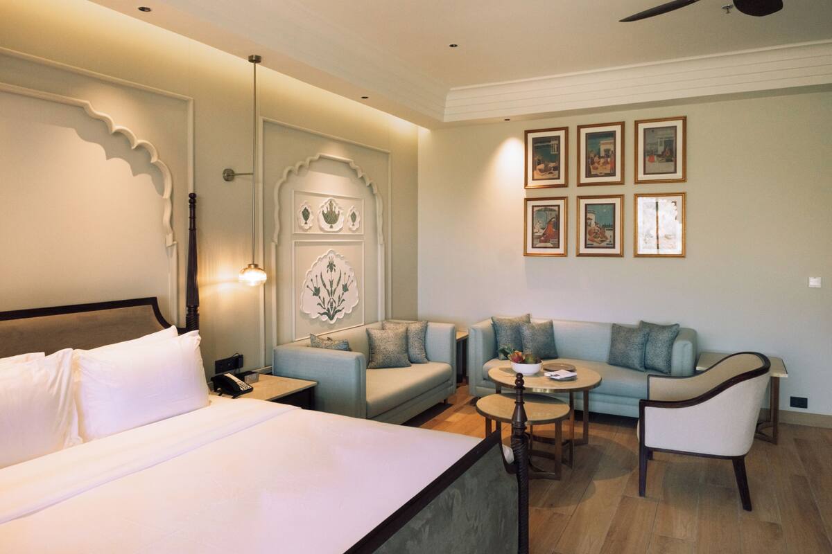 Habitación de hotel con cama, sofá, mesita y una silla. Se observan cuadros enmarcados en la pared y un panel decorativo.
