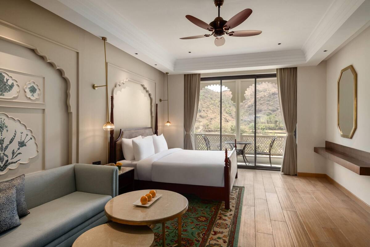 Habitación de hotel con cama, sofá, una mesa redonda con naranjas, ventilador de techo y balcón con vistas.