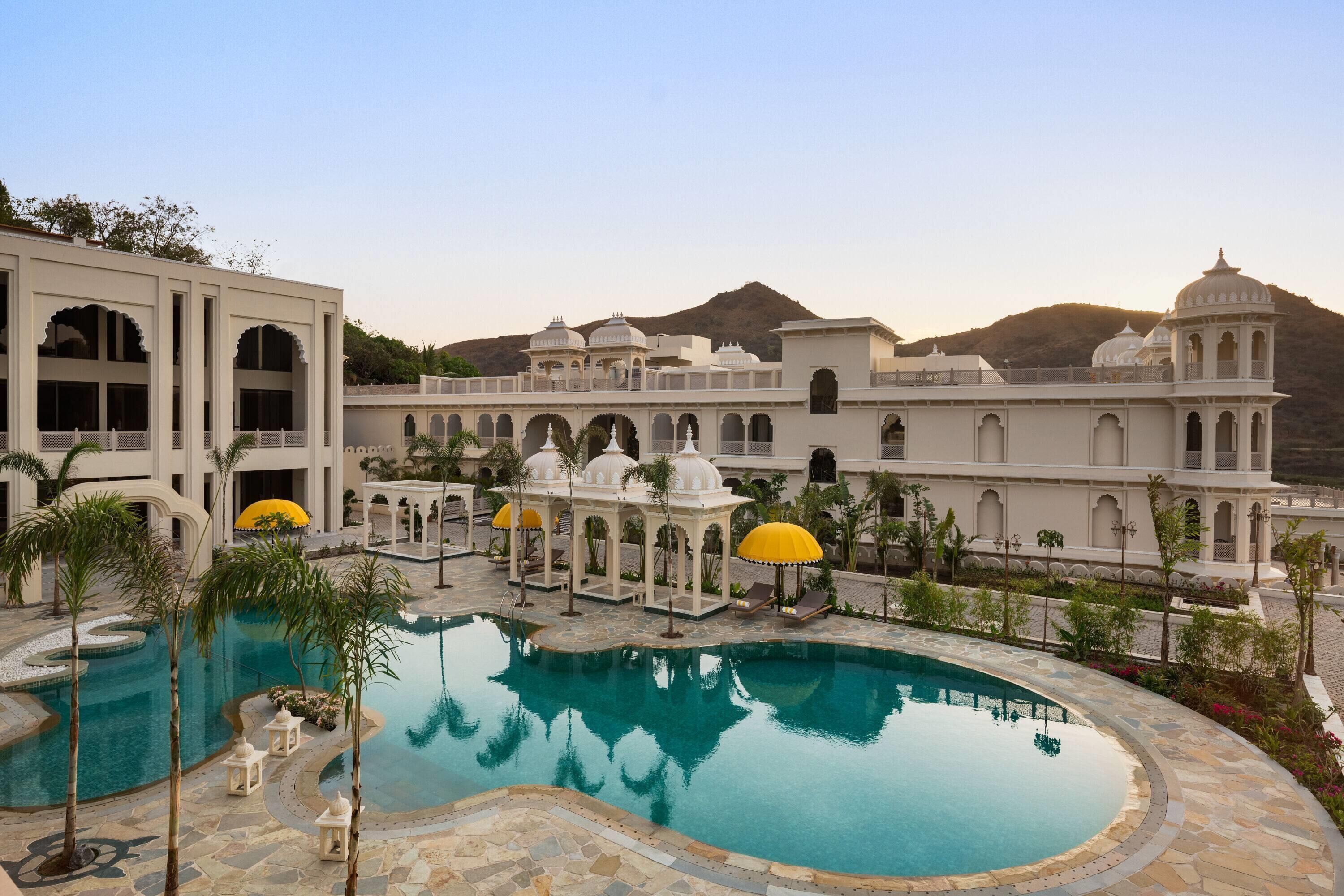 A pool surrounded by a white building with arches and domes, palm trees, and a mountainous backdrop.