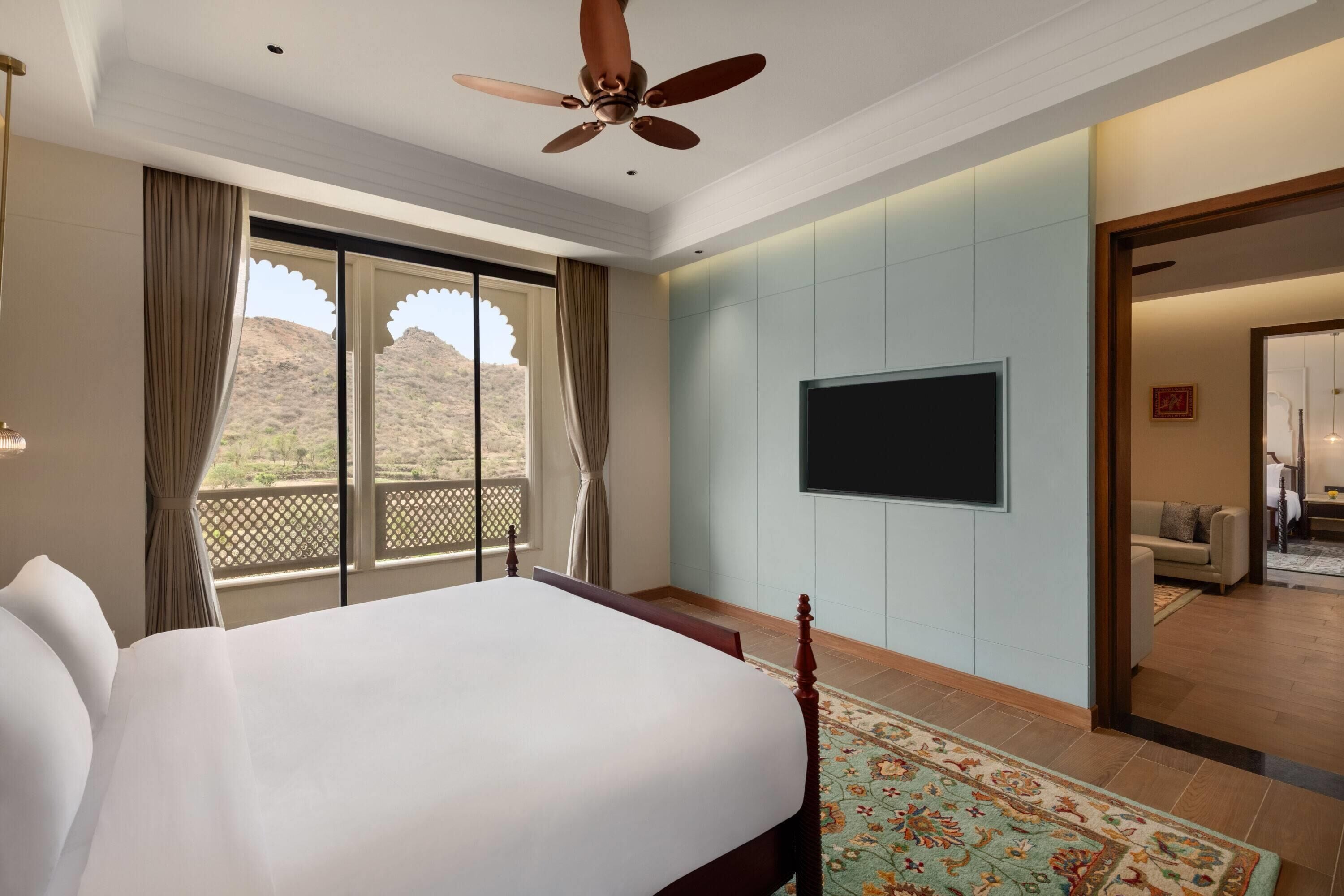 A hotel room with a large bed, a ceiling fan, a TV, and a balcony with curtains.