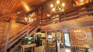 A rustic cabin interior with wooden walls, a staircase, a dining area, and a kitchen. - Luxury Secluded Lodge with a View | Hot Tub•Games (Logan)