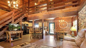 A rustic cabin interior with a wooden staircase, a dining area with chairs, a sofa, and a mounted television. - Luxury Secluded Lodge with a View | Hot Tub•Games (Logan)