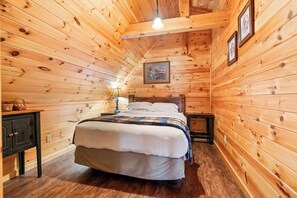 A wooden cabin bedroom with a bed, nightstands, a lamp, and framed pictures on the walls. - Cozy Secluded Lodge with a View | Hot Tub•Games (Logan)