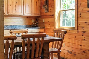 A wooden cabin interior with a dining table and chairs, a window with a view of greenery, and a countertop with a bottle. - Cozy Secluded Lodge with a View | Hot Tub•Games (Logan)