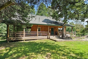 A wooden cabin with a porch, surrounded by trees and a grassy area.