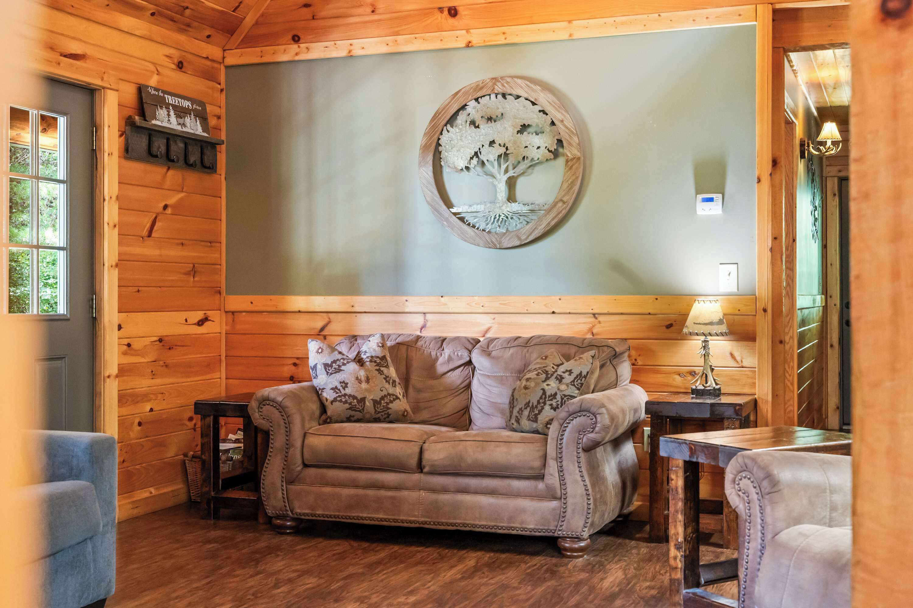 A cozy cabin interior with a wooden wall, a round mirror with a tree design, a brown leather sofa, and a small table with a lamp.