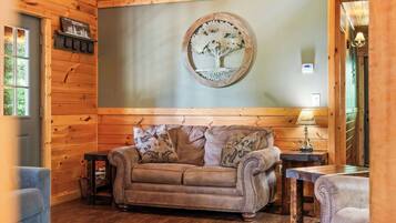 A cozy cabin interior with a wooden wall, a round mirror with a tree design, a brown leather sofa, and a small table with a lamp.