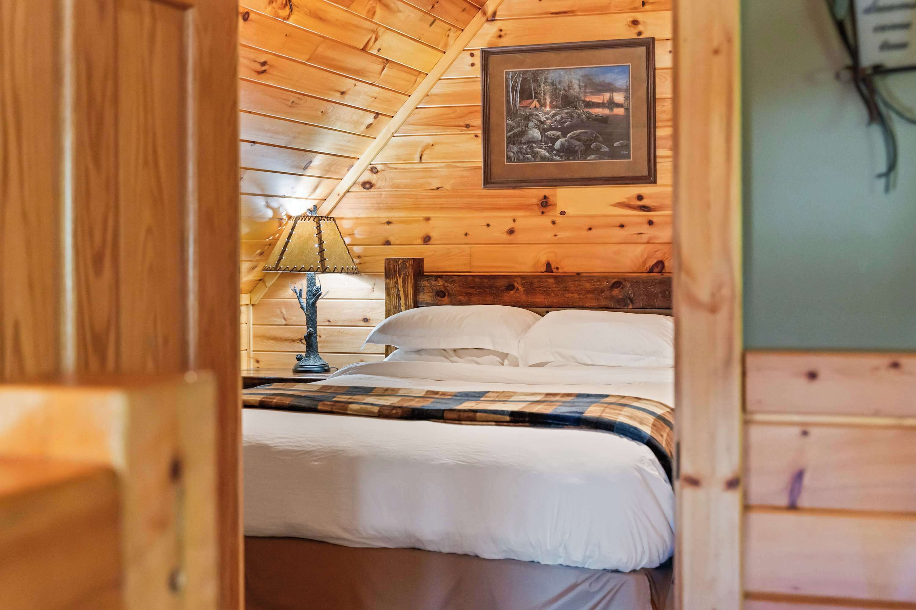 A wooden cabin room with a bed, a nightstand, a lamp, and a framed picture on the wall.