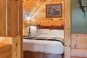 Ein Zimmer einer Holzhütte mit einem Bett, einem Nachttisch, einer Lampe und einem gerahmten Bild an der Wand.